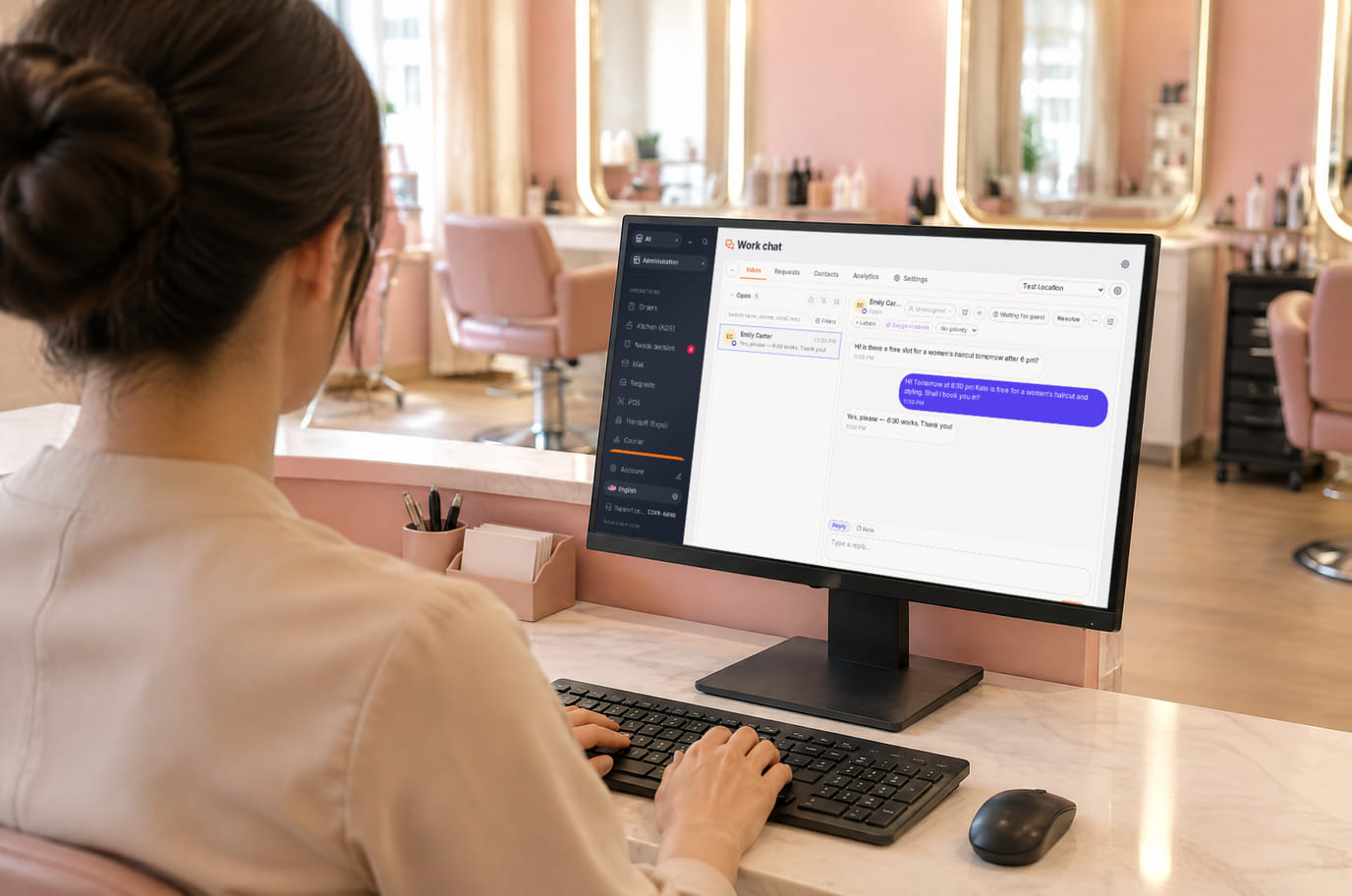 A salon receptionist at the front desk answers website visitors in the chat inbox on a monitor